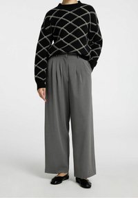 Svart stickad tröja med vitt diamantmönster, kombinerad med grå vida byxor med veck. Svarta platta skor fulländar outfiten.