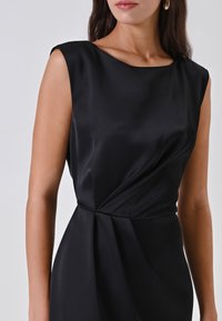 Donna in un vestito senza maniche di satin nero con dettagli drappeggiati in vita e orecchini a cerchio dorati su uno sfondo chiaro e semplice.