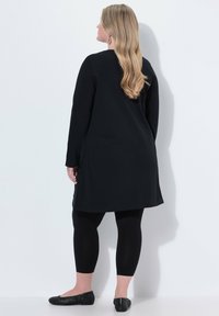 Schwarzes langärmliges Tunika mit seitlichen Schlitzen, kombiniert mit schwarzen Leggings und flachen Schuhen mit einem geflochtenen Muster. Das Haar reicht bis zur Mitte des Rückens.