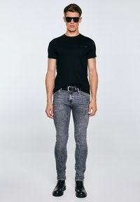 Vyras dėvi juodus saulės akinius, prigludusią juodą marškinėlę, pilkus rūgštinio plovimo džinsus su juodu diržu ir juodus odinius batus, stovintis priešais paprastą foną.