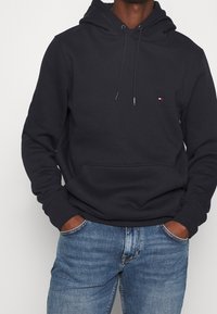 Haut à capuche marine en mélange de coton, avec une poche avant et des cordons de réglage. Petite logo sur la poitrine. Associé à un jean bleu.