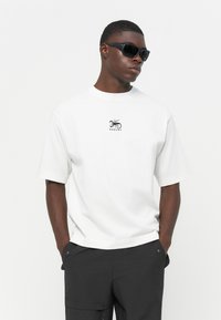 Hvid bomulds t-shirt med et sort trykt grafisk motiv på brystet, parret med sorte bukser. Model med sorte solbriller.