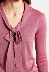 Różowy sweter dzianinowy z szerokim kołnierzem i dodatkowym detalem w postaci kokardy przy dekolcie, z długimi rękawami i gładką fakturą.