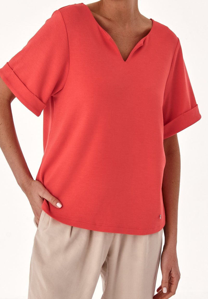 Haut corail à col en V avec manches courtes retroussées, confectionné en tissu lisse. Associé à un pantalon beige clair, avec un détail subtil de logo.