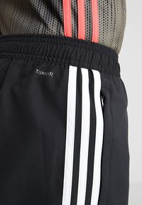 Shorts sportivi neri con dettagli a tre righe bianche, vita elastica e vista parziale di una maglia sportiva in rete con righe rosse.