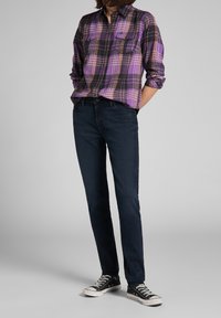 Chemise en flanelle à carreaux violets et noirs avec deux poches poitrine, assortie à un jean bleu foncé et des baskets noires avec des semelles en caoutchouc blanches.
