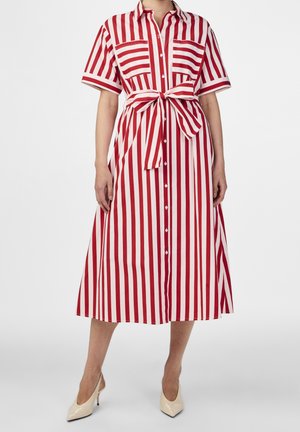 Vrouw draagt een midi overhemdjurk met verticale rood-witte strepen, korte mouwen, knopen aan de voorkant, borstzakken, tailleband en beige hakken.