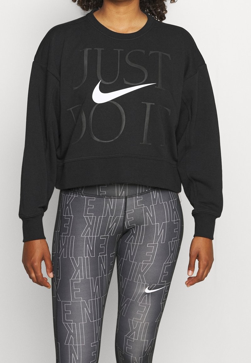 Personne portant un sweat court noir avec le texte "JUST DO IT" et le logo Nike blanc, associé à des leggings gris foncé avec le texte Nike répété.