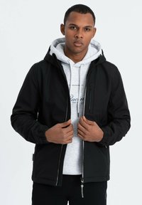 Giacca nera con zip e cappuccio, dotata di polsini elastici e una tasca sul petto. Indossata sopra un hoodie grigio chiaro con un motivo stampato.