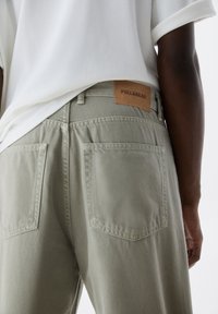 Personne portant un jean gris clair avec des poches arrière et une étiquette marron Pull&Bear, assorti à une chemise blanche aux manches retroussées.