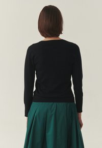 Pull noir à manches longues avec une coupe ajustée et des poignets côtelés, porté par-dessus une jupe plissée verte, vu de dos.