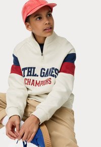 Sudadera color crema con rayas azules y rojas, cuello con cremallera y texto "ATHL. GAMES CHAMPIONS"; pantalones beige; zapatos azules; gorra rosa.