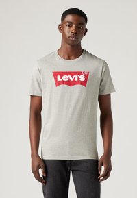 Siva pamučna majica s crvenim Levi's logom u središtu prsnog dijela. Ima kratke rukave i dizajn s okruglim izrezom.