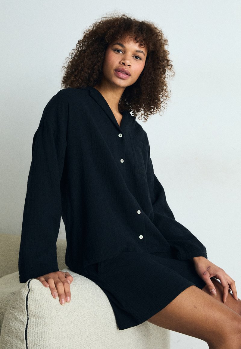Schwarzes Leinen-Hemd mit Knöpfen und Kragen, kombiniert mit einem passenden Rock. Weicher, strukturierter Stoff. Model sitzt auf einem hellen Sofa.