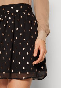 Jupe noire avec un motif de pois dorés, dotée d'une taille cintrée et d'un design en couches. Le tissu présente un aspect léger et texturé.