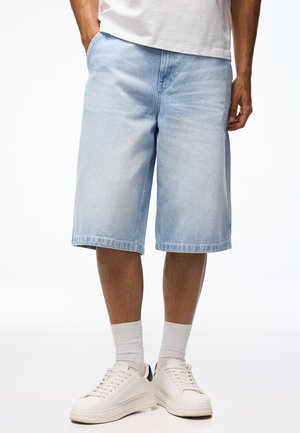 Férfi világoskék farmer térdnadrágot, fehér zoknit, fehér sportcipőt és fehér inget visel, a kezét zsebre téve.