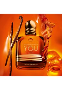Pomarańczowa szklana buteleczka perfum z złotą nakrętką, z napisem "Stronger With You Amber" oraz akcentem strąka wanilii obok.