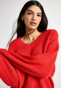 Červený svetr s výstřihem do V z měkkého, nadýchaného úpletu s volnými rukávy, vrstvenou texturou a volným střihem, s hladkým povrchem a minimálním zdobením.