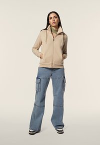 Felpa con zip beige con lati testurizzati, abbinata a jeans cargo blu a gamba larga con grandi tasche e cuciture a contrasto. Scarpe bianco e nere.