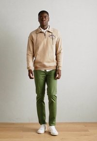 Beige sweatshirt with a half-zip and embroidered design, paired with green pants and white sneakers, standing against a neutral background.