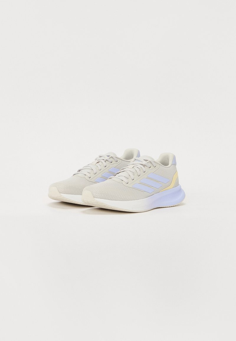 Zapatillas deportivas de color gris claro con parte superior de malla transpirable, acentos en color lila, logo de tres rayas y una suela blanca acolchada.
