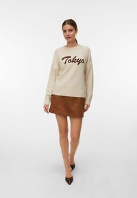 Vrouw met een paardenstaart, gekleed in een beige sweater met de tekst "Tokyo", een bruin suède mini-jurk en zwarte puntige flats, staande tegen een witte achtergrond.