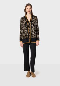 Svart kavaj med guldprickar, öppen front och kontrasterande orange kant. Bärs över en svart topp och matchas med svarta byxor och bruna skor.