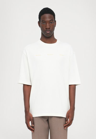 White cotton t-shirt with oversized fit, short sleeves, and round neck. Three lines of text in yellow across the chest. Light texture.