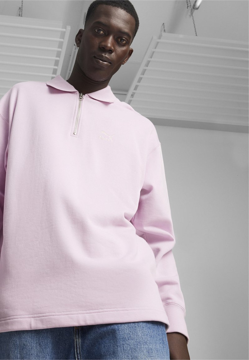 Roze sweatshirt met een kwartlengte rits en een kraag, met een klein wit logo. De stof lijkt zacht en textuur te hebben.