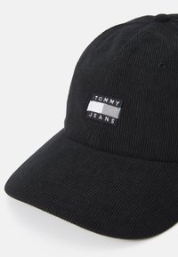 Fekete kordbársony sapka ívelt szélekkel, elöl egy téglalap alakú folttal, amelyen fehér és szürke színnel szerepel a "TOMMY JEANS" felirat.