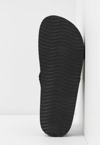Semelle noire de sandale avec un motif ondulé, debout verticalement contre un mur d'angle blanc et un sol.