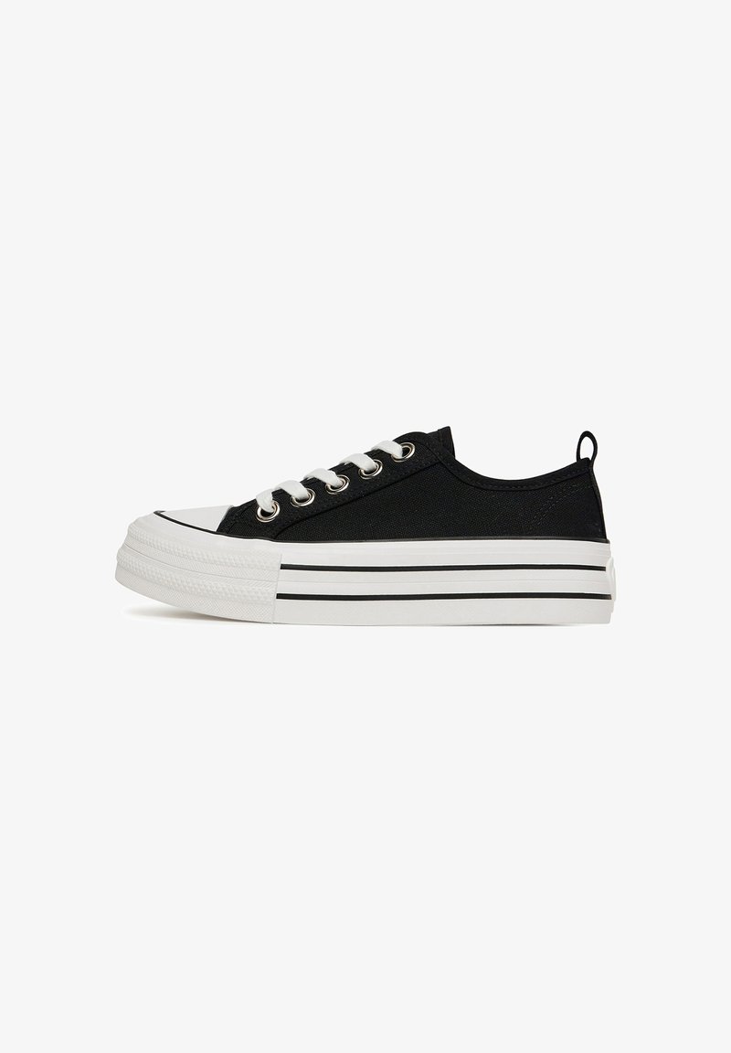 Zapatillas de lona negras con suelas de goma blancas, que presentan detalles a rayas negras y blancas, puntera redonda y cordones blancos.