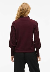 Burgundy tröja med texturerade, puffiga ärmar och ribbad krage. Slät material på kroppen, kontrasterande stickmönster på ärmarna.