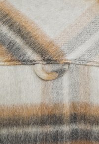 Ulltyg med ett rutmönster i beige, bruna och grå nyanser. Har en rund knappdetalj och en mjuk textur.