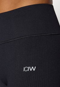 Zwarte geribde legging met een hoge tailleband, voorzien van een witte "ICIW"-logo aan de linksonderzijde. Gladde textuur, figuurvormend ontwerp.