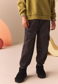 Pantaloni cargo grigio scuro con polsini elasticizzati e tasche, abbinati a stivaletti neri in suede. Il modello indossa una felpa verde con polsini a righe.