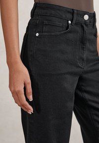 Close-up of a person wearing high-waisted black denim jeans with visible front pockets and a hand relaxed by their side.