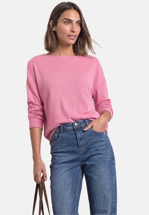 Kvinde iført pink langærmet sweater og blå jeans, holder brune taskestropper i den ene hånd, står mod en ensfarvet baggrund.