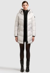 Lysegrå pufferjakke med hætte, med lynlås foran, to sidelommer og et quiltet design. Båret over en sort kjole og strømpebukser.