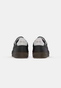 Paire de baskets basses noires avec collets de talon blancs et semelles marron, logo doré "BOSS" au dos de chaque chaussure.