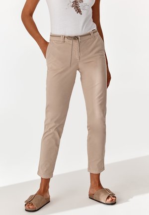 Kobieta w beżowych wąskich spodniach z troczkiem w pasie, białym t-shircie z nadrukiem i beżowych klapkach na tle jednolitej białej ściany.