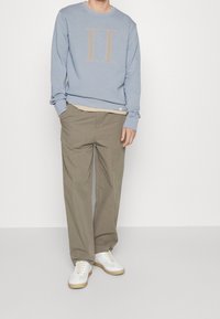 Felpa blu chiaro con logo beige "II", abbinata a pantaloni chinos color khaki e sneakers bianche, dotate di lacci e suola texturizzata.