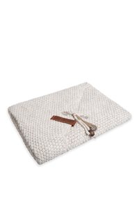 Gebreide crèmekleurige deken met een gestructureerd patroon, gevouwen met een leren label en decoratieve strikken met metalen accenten.