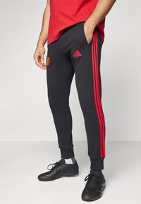 Calças de treino pretas com riscas laterais vermelhas, apresentando um emblema do Manchester United e o logótipo da Adidas, feitas de um tecido macio e texturizado.