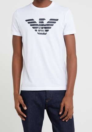 T-shirt en coton blanc avec un logo d'aigle rayé noir sur le devant. Col rond, manches courtes et coupe décontractée.