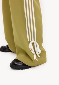 Olijfgroene wijde broek met witte zijkanten en omstrikbare zomen, gecombineerd met glanzend zwarte schoenen met een geribbelde zool.