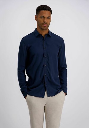 Mann steht vor einfachem Hintergrund, trägt ein dunkelblaues Hemd mit Knöpfen und hellbeige Hosen, die Hände in den Taschen.