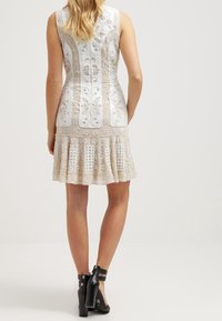 Robe sans manches blanche avec des accents beiges, ornée de broderies florales, d'un ourlet évasé et de motifs texturés ; associée à des bottes à talons noires.