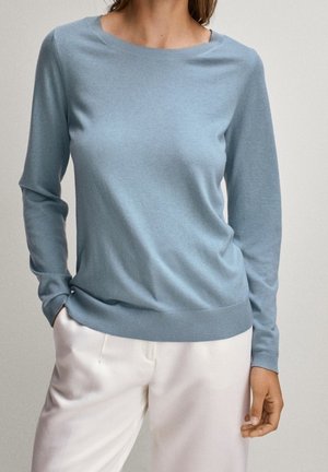 Femme portant un pull bleu clair à manches longues et un pantalon blanc, debout avec une main dans la poche devant un fond neutre.