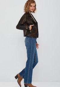 Giacca di pelle marrone con un design aderente, tasche con zip, texture consumata, abbinata a jeans in denim blu e stivali con tacco marroni.